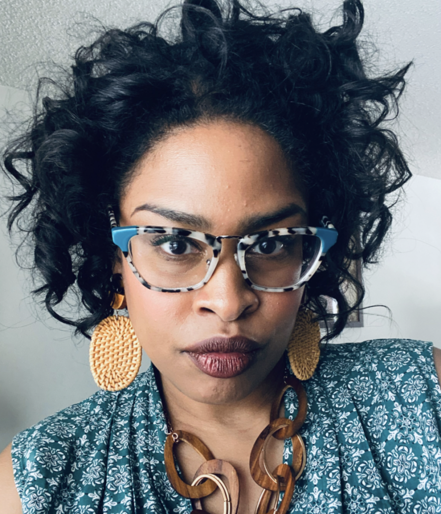 Allison wearing bold glasses and jewelry, with a blue patterned top, in front of a white background.
