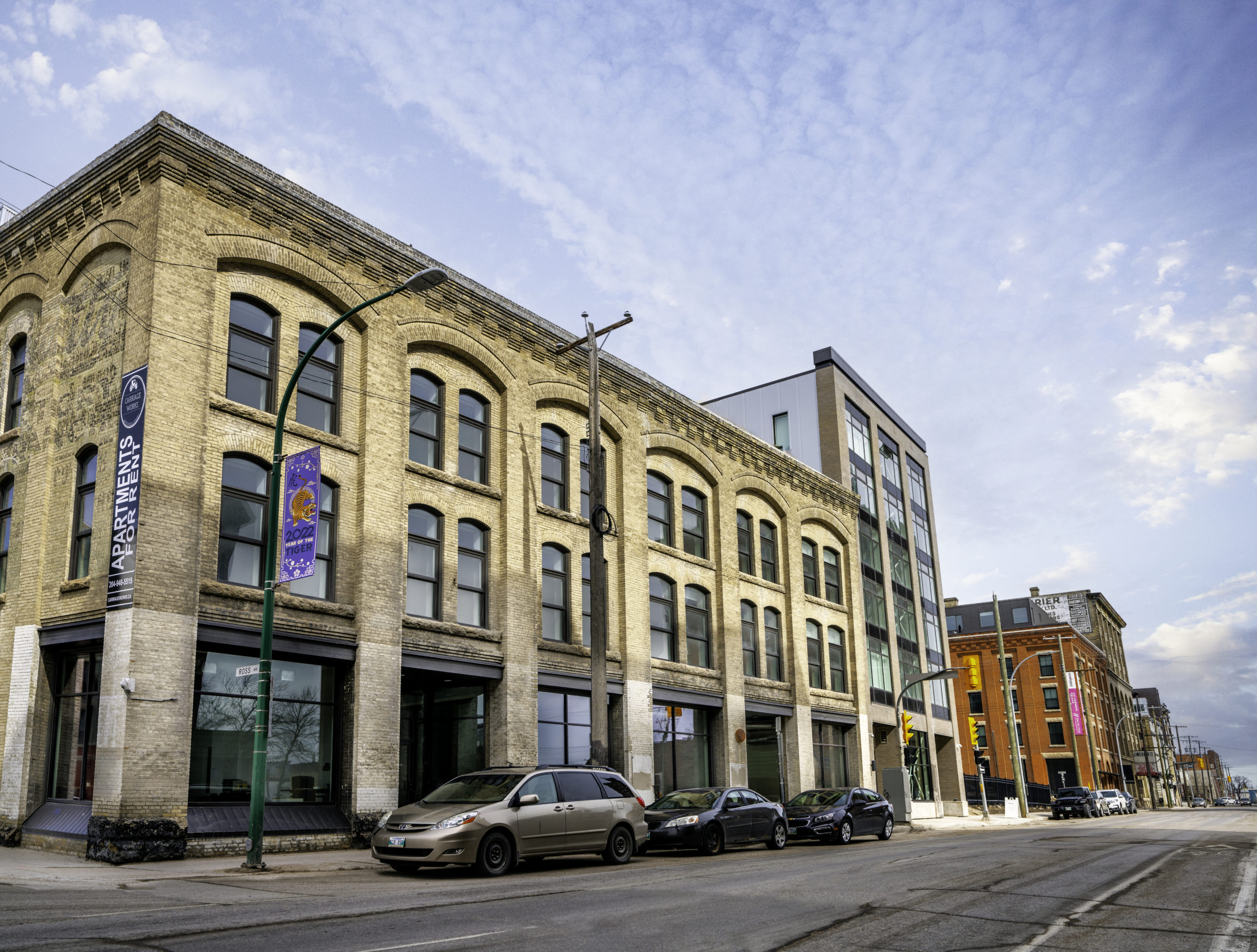 A view of the block on which the gallery is located. The building is made of brick with large windows on the main floor. There is a tall modern building at the end with many windows. 3 cars are parked in front of the building. The sun is shining. 