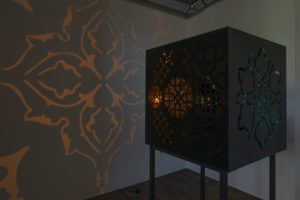 An installation photograph of Zahra Baseri's piece Petrocubical. The piece is a black box with designs cut out of the sides, with turquoise and amber lights placed inside of it. The light shines through the cut out designs and projects patterns on the walls around it.