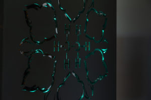 A close up photograph of Zahra Baseri's piece Petrocubical. The piece is a black box with designs cut out of the sides, with turquoise and amber lights placed inside of it.